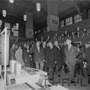 grupa mężczyzn, część w płaszczach, i kilka kobiet ogląda ekspozycję w hali targowej