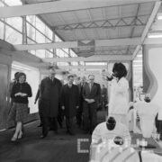 grupa mężczyzn w płaszczach, z lewej kobieta w hali wystawowej ogląda stoisko z odzieżą prezentowaną na manekinach