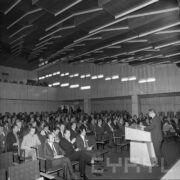 dużo osób siedzi na krzesłach ustawionych w lekkim układzie amfiteatralnym, z prawej mównica, przy niej stoi mężczyzna, w głębi balkon, na nim widoczni ludzie