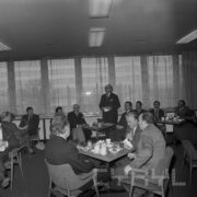 grupy mężczyzn siedzących przy stolikach, na których przygotowano poczęstunek, pośrodku jeden mężczyzna stoi i przemawia