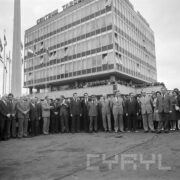 na pierwszym planie tłum ludzi, większość mężczyzn, za nim fragment wysokiego budynku, na balkonie, na pierwszym piętrze stoją ludzie