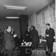 grupa mężczyzn stoi, przy nich stoliki, na których przygotowano poczęstunek, część z mężczyzn trzyma w rękach kieliszki do szampana, pośrodku dwóch mężczyzn podaje sobie dłonie