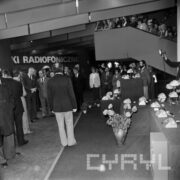 hala targowa, duża grupa mężczyzn i kobiet stoi przy stoisku z aparatami telefonicznymi, w głębi na schodach duża grupa ludzi