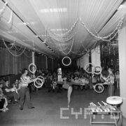 na pierwszym planie dwóch mężczyzn stojących do siebie twarzami żagluje kółkami, pomiędzy nimi na jednej nodze stoi kobieta, w głębi dużej sali widownia złożona z dzieci, kobiet i mężczyzn, z sufitu zwisają serpentyny