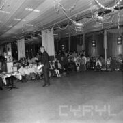 duża sala, z sufitu zwisają serpentyny, w głębi duża grupa dzieci siedzi na krzesłach na kształt widowni, przed nimi stoi starszy mężczyzna