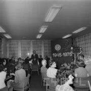 grupy kobiet i kliku mężczyzn siedzi przy stolikach, na których przygotowano poczęstunek, w głębi stoi mężczyzna i przemawia