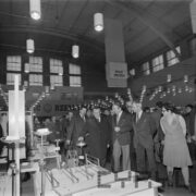 grupa mężczyzn w płaszczach, część w garniturach, z prawej kobieta w hali wystawowej ogląda stoiska z towarami wystawowymi