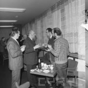 czterech mężczyzn zwróconych do siebie stoi przy małym stoliku, na którym przygotowano poczęstunek, w rękach trzymają kieliszki