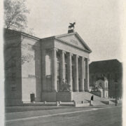 monumentalny budynek z kolumnadą, do budynku prowadzą schody, po obu ich stronach stoją kamienne rzeźby lwów, na których siedzą ludzie; na dachu budynku umiejscowiona została figura pegaza