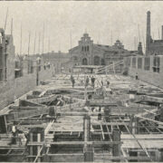 grupa ludzi stojąca i pracująca na drewnianych rusztowaniach obok murów z cegieł; w głębi budynek z cegieł