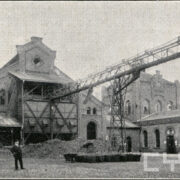 ceglany budynek z kratownicą służącą do transportu węgla; w głębi zabudowania i kominy