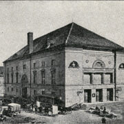 duży budynek na placu, przed budynkiem dorożki i grupa ludzi