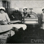 czwórka dziewcząt w czasie prasowania; dwie z nich prasują przy pomocy żelazek; dwie kolejne obsługują magiel