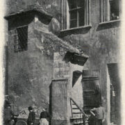 wejście do kamienicy ze zniszczoną elewacją, przed budynkiem stoi trzech mężczyzn i kobieta; jeden z mężczyzn opiera rower o ścianę