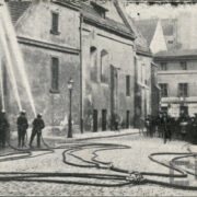 pięciu mężczyzn w strojach strażackich i hełmach polewający wodą z węży duży budynek z cegieł; po drugiej stronie stoi grupa gapiów