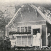 grupka dzieci z mężczyzną w kapeluszu (prawdopodobnie z nauczycielem) przed drewnianym domkiem w stylu szwajcarskim, w drzwiach którego stoi dwójka sprzedawców w białych fartuchach