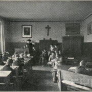 grupa uczniów siedzących w drewnianych ławkach; widoczne meble, a na ścianach obraz i trzy mapy oraz krzyż
