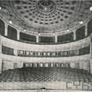 rzędy krzeseł w pięknie zdobionej sali; widoczne balkony i ozdobne sklepienie pomieszczenia